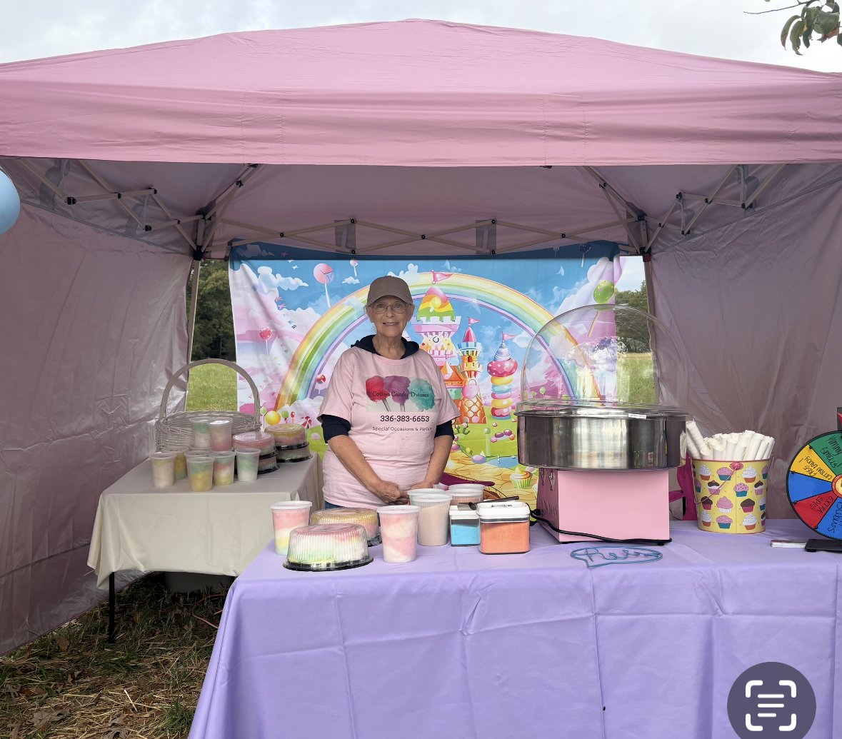Carol Greve, owner of Cotton Candy Dreams, at an outdoor event booth with cotton candy machine and colorful rainbow backdrop